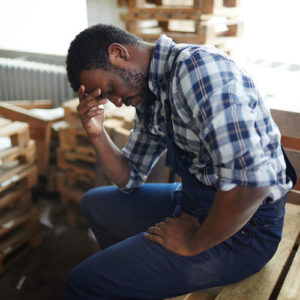 distressed working class employee buries his face in his hand as he sits atop a stack of wooden pallets