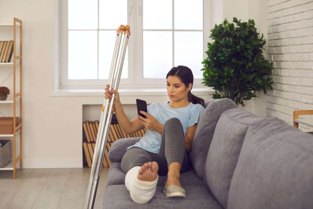 Woman with a broken leg sitting on the couch holding crutches in one hand and her phone in the other